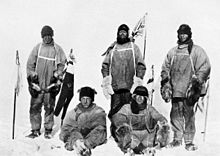Five men(three standing, two sitting on the icy ground) in heavy polar clothing. All look exhausted and unhappy. The standing men are carrying flagstaffs and a Union flag flies from a mast in the background. Scott's party at the South Pole. Left to right: Oates; Bowers; Scott; Wilson; Evans